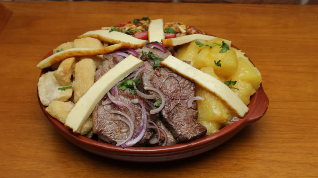 Travessa de barro com ribação fatiada e acebolada com cebola roxa, acompanhada de macaxeira frita, macaxeira cozida e tiras de queijo coalho grelhado.